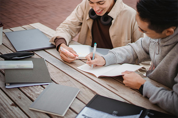 two individuals sitting at a table writing in a notebook. Pexels - Armin Rimoldi